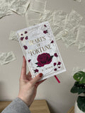 Hand holding a book titled 'Hearts of Fortune' by E. Winn with rose design on a surface with scattered pages on the wall behind the image.