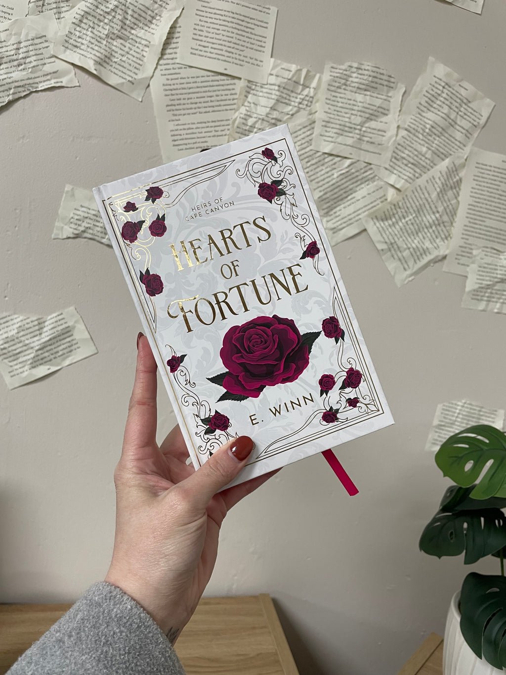 Hand holding a book titled 'Hearts of Fortune' by E. Winn with rose design on a surface with scattered pages on the wall behind the image.