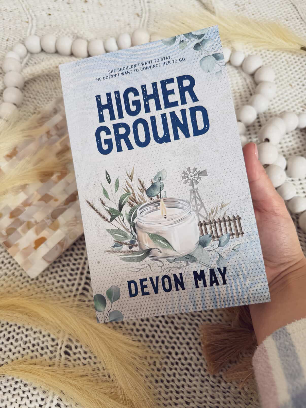 Book titled 'Higher Ground' by Devon May held by a hand on a textured surface.